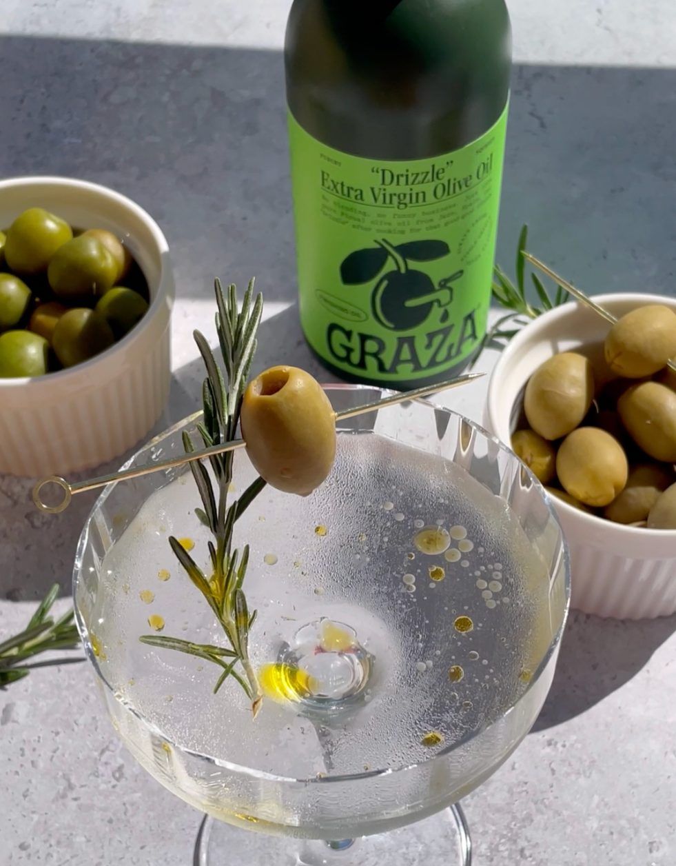 A bottle of “Drizzle” behind a martini with olives